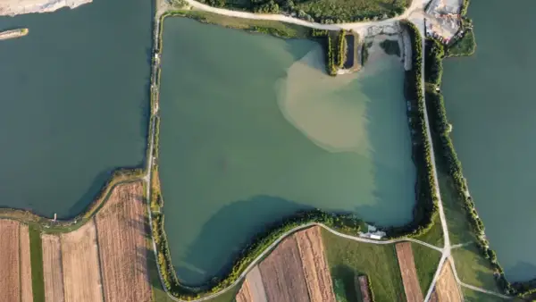 Widok z lotu ptaka na zielone oczko wodne, otoczone polami i drogami. Idealne miejsce na spokojne łowisko.